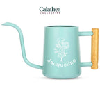A turquoise metal watering can with long gooseneck spout and wood handle with a white flower bouquet design and 'Jacqueline' below the bouquet in white permanent vinyl.