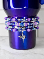 A deep purple tumbler adorned with multiple beaded bracelets in shades of purple, blue, and silver. A silver charm with a starburst design hangs from the bracelets. The tumbler has a gradient finish, transitioning from dark to lighter purple.