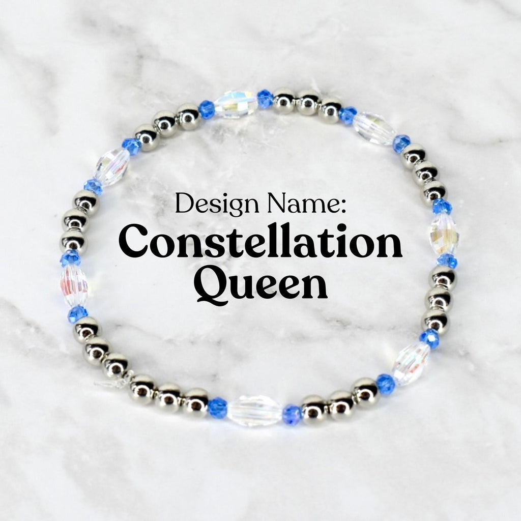A beaded bracelet with the text "Design Name: Constellation Queen". The bracelet features silver-colored beads, blue beads, and clear, iridescent crystal beads. The bracelet is circular and sits on a white and gray marble surface.