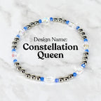 A beaded bracelet with the text "Design Name: Constellation Queen". The bracelet features silver-colored beads, blue beads, and clear, iridescent crystal beads. The bracelet is circular and sits on a white and gray marble surface.