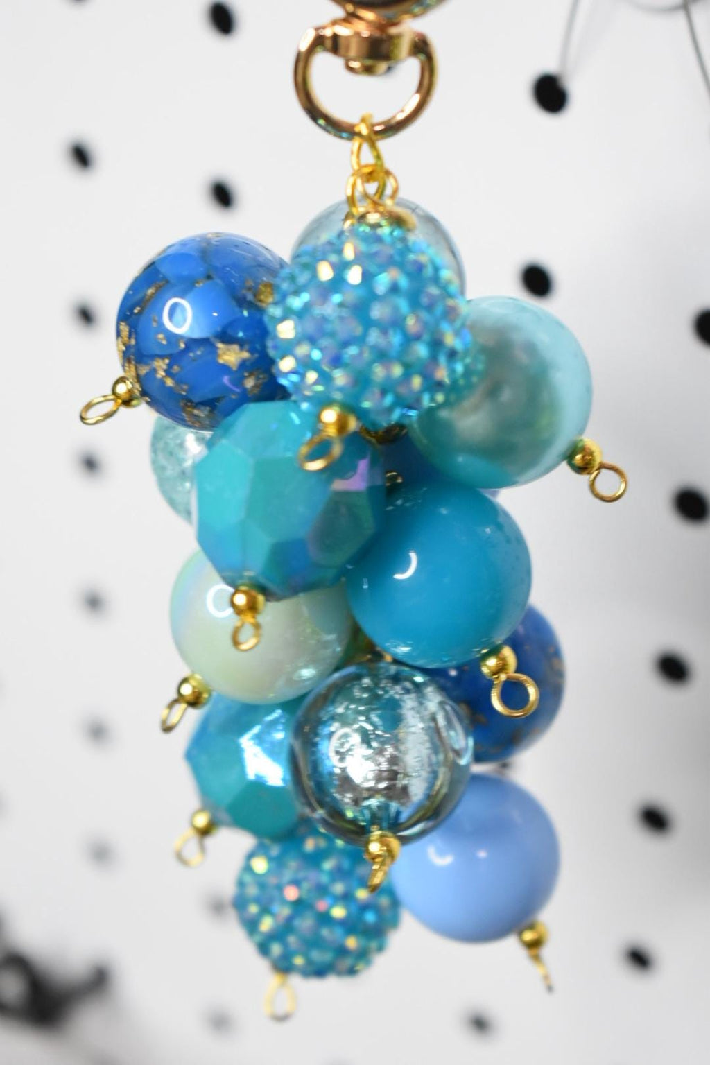 A cluster of blue and turquoise beads with gold-colored accents. The beads vary in size, shape, and texture, including faceted, smooth, and glittery surfaces. A gold-colored clasp is at the top.