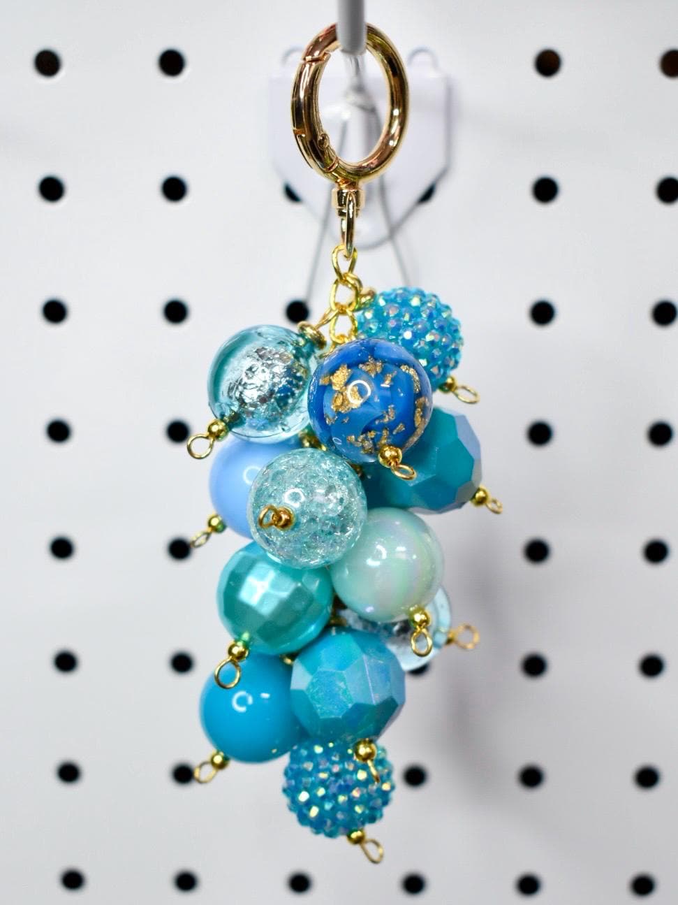 A cluster of blue and turquoise beaded charms hangs from a gold-colored keychain ring. The beads vary in size, shape, and texture, including faceted, glittery, and textured designs. The keychain is suspended against a white background with black dots.