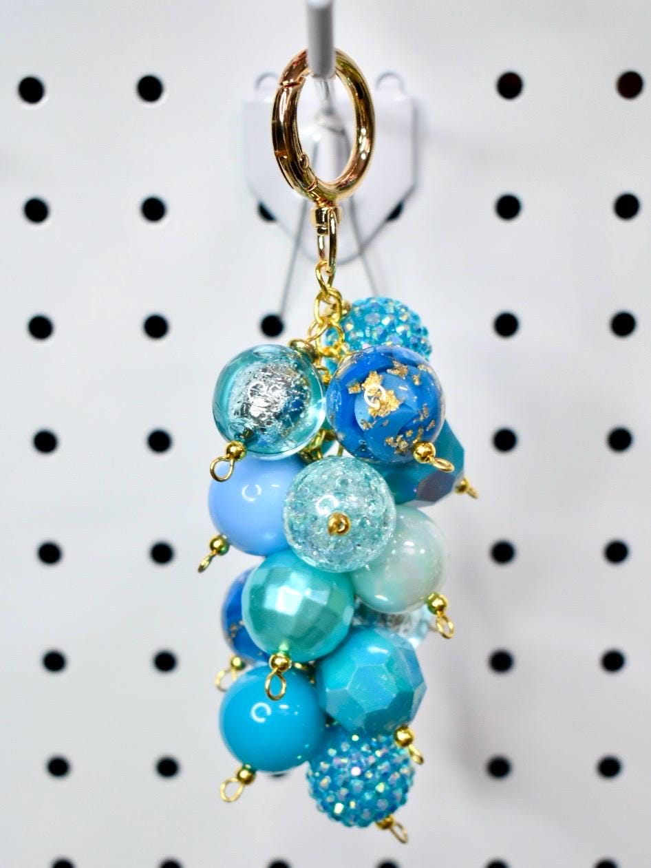 A cluster of blue beaded charms hangs from a gold-colored keychain clasp. The beads vary in shades of blue, including light blue, turquoise, and navy. Some beads have glitter or gold leaf accents. The keychain is suspended against a white background.