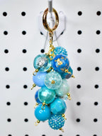 A cluster of blue beaded charms hangs from a gold-colored keychain clasp. The beads vary in shades of blue, including light blue, turquoise, and navy. Some beads have glitter or gold leaf accents. The keychain is suspended against a white background.