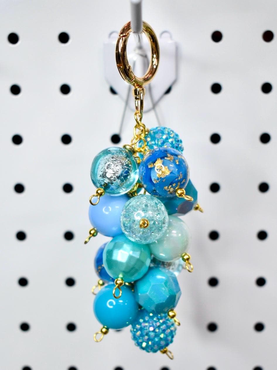 A cluster of blue beaded charms hangs from a gold-colored keychain clasp. The beads vary in shades of blue, including light blue, turquoise, and navy. Some beads have glitter or gold leaf accents. The keychain is suspended against a white background.