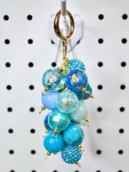 A cluster of blue beaded charms hangs from a gold-colored keychain clasp. The beads vary in shades of blue, including light blue, turquoise, and navy. Some beads have glitter or gold leaf accents. The keychain is suspended against a white background.