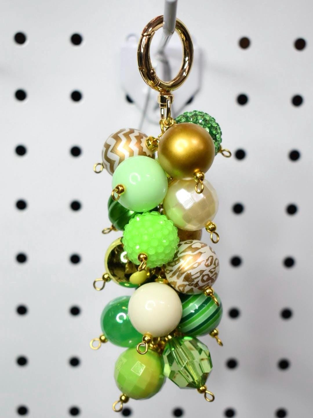 A beaded keychain with a gold clasp and a cluster of green, gold, and cream-colored beads. The beads vary in size, shape, and texture, including solid, patterned, and faceted designs. The keychain is hanging against a white background.