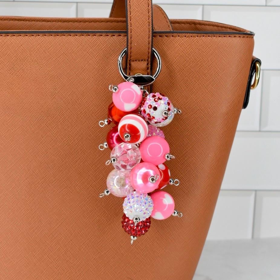 A pink and red beaded keychain with a variety of bead sizes and textures. The keychain is attached to a brown leather handbag. The beads include solid pink, striped, and crystal-covered beads.
