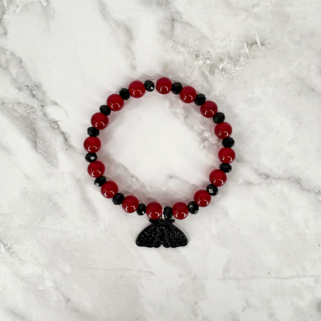 A circular beaded bracelet featuring alternating smooth red spherical beads and smaller faceted black beads. A black moth-shaped charm hangs from the bottom of the bracelet, centered among the beads, creating a distinct focal point.