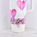 A white tumbler with pink and purple heart designs, adorned with multiple beaded bracelets in shades of pink, purple, and silver. A silver heart charm dangles from the bracelets, adding a touch of elegance.
