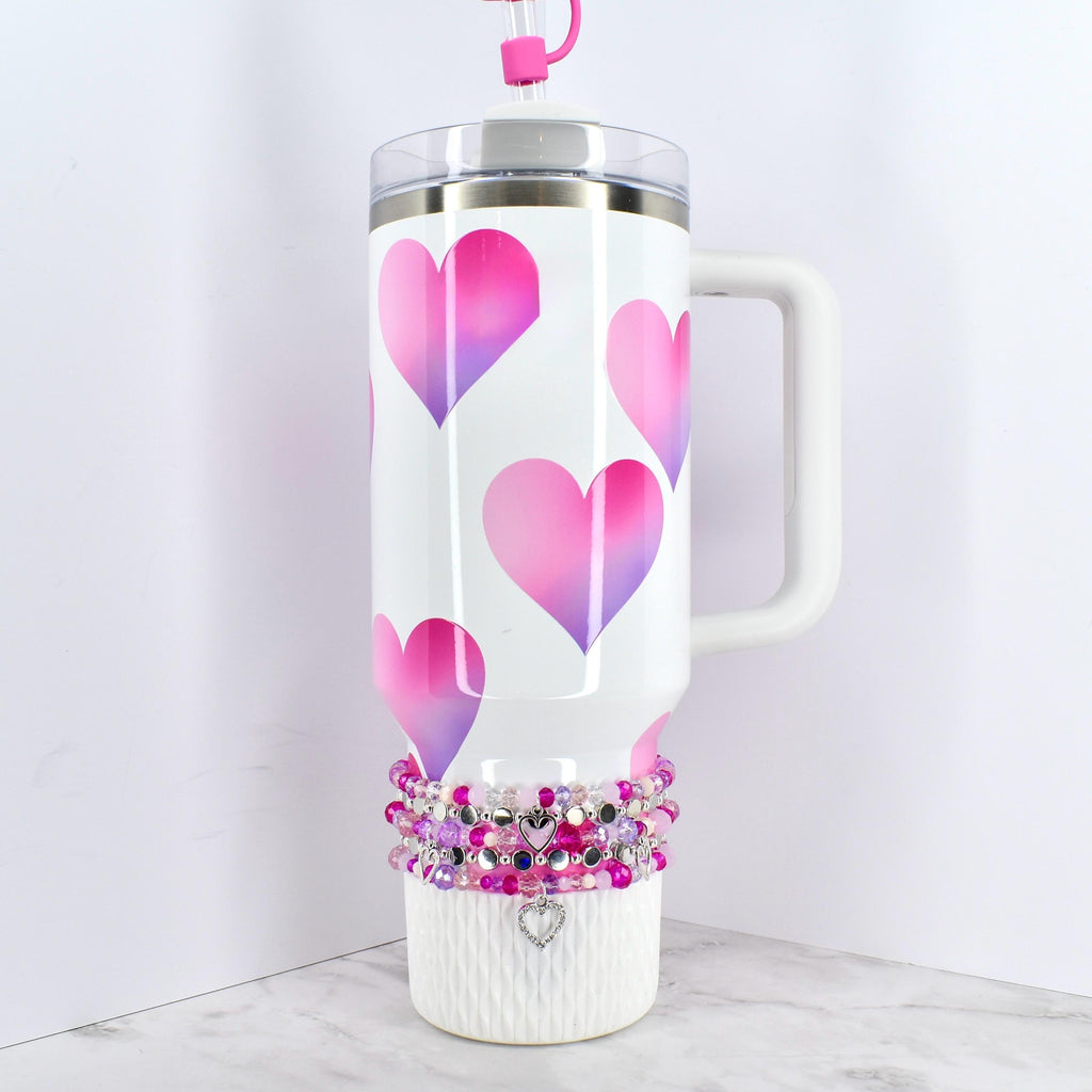 White insulated tumbler with a handle, featuring a pattern of pink and purple gradient hearts. The tumbler has a clear lid and a pink straw. A beaded bracelet with a heart charm decorates the base.