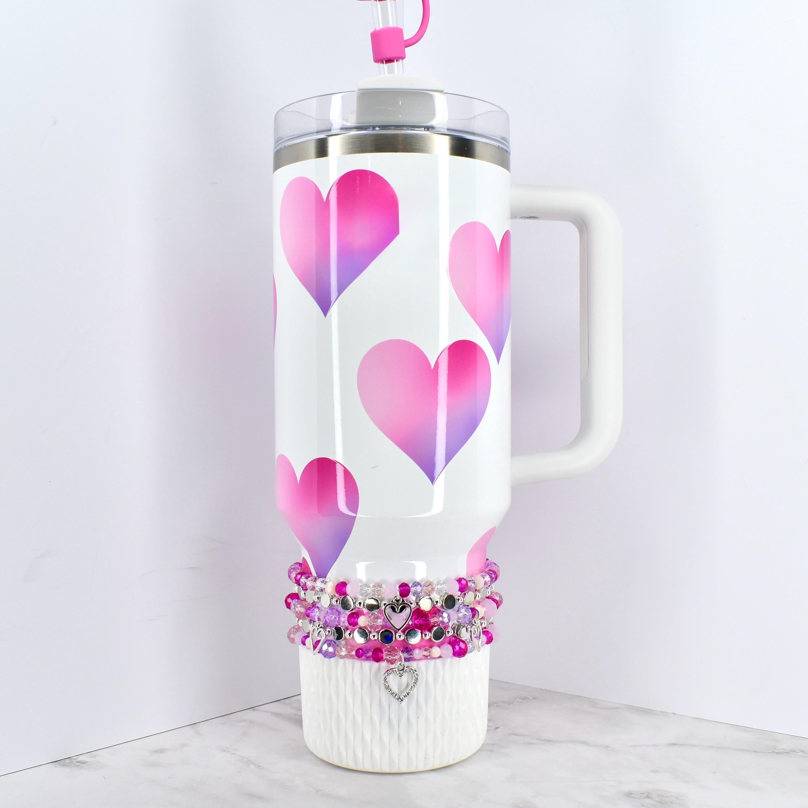 White insulated tumbler with a handle, featuring a pattern of pink and purple gradient hearts. The tumbler has a clear lid and a pink straw. A beaded bracelet with a heart charm decorates the base.