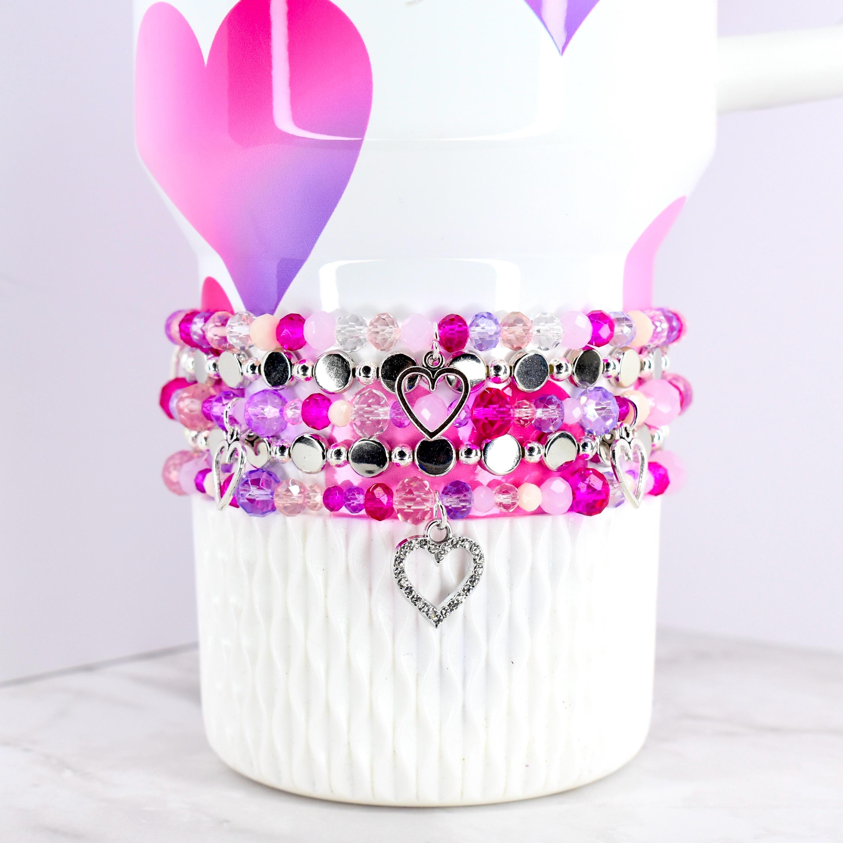 Stack of beaded bracelets in shades of pink, purple, and clear, with silver heart charms. The bracelets are displayed on a white mug decorated with pink and purple hearts.