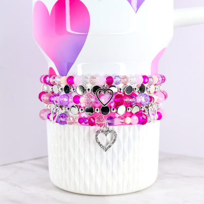 Stack of beaded bracelets in shades of pink, purple, and clear, with silver heart charms. The bracelets are displayed on a white mug decorated with pink and purple hearts.