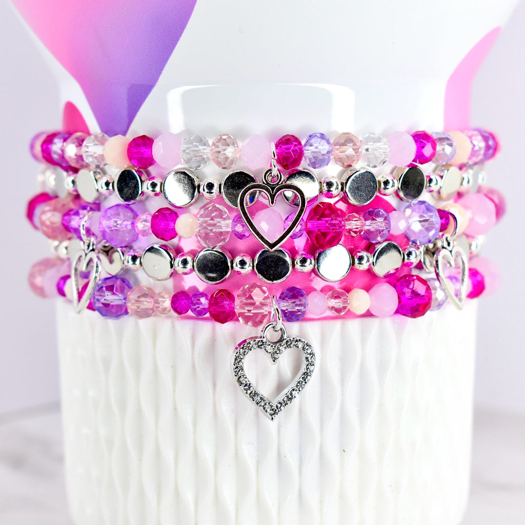 Stack of beaded bracelets in shades of pink, purple, and clear beads. Each bracelet has silver accents and a heart charm. The bracelets are displayed on a white textured surface.