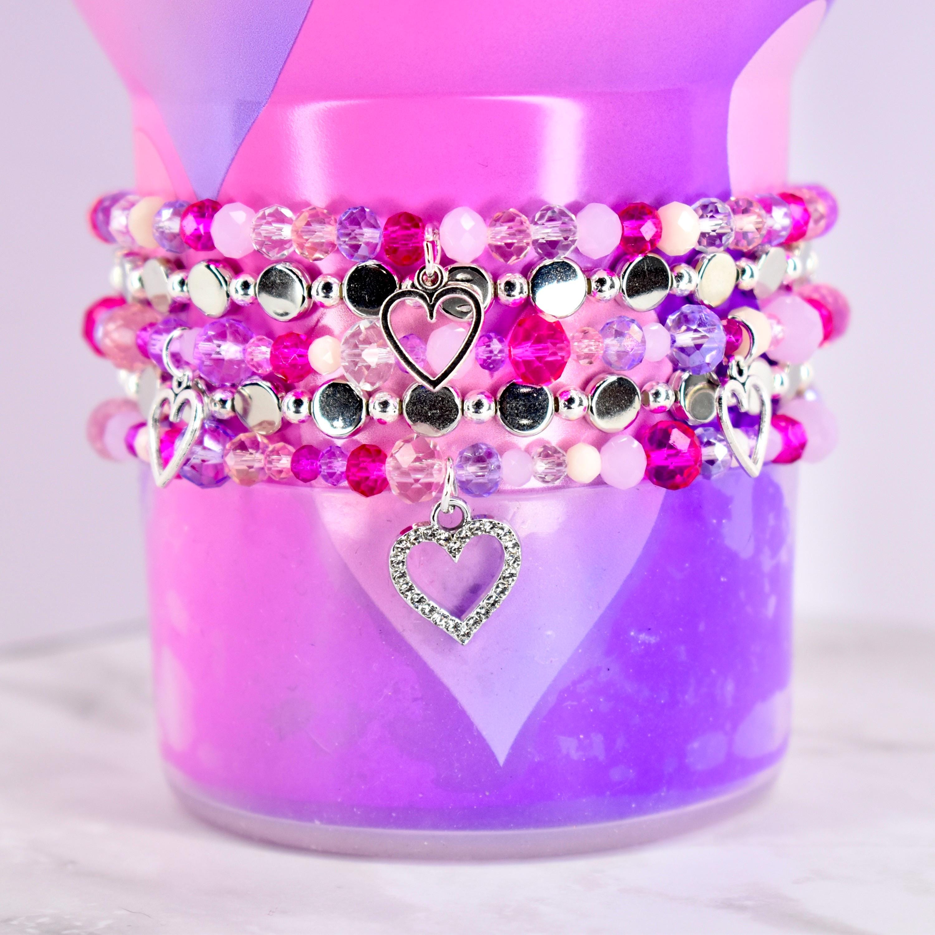 Stack of beaded bracelets in shades of pink, purple, and silver. Each bracelet features a variety of beads and a heart charm. The bracelets are displayed on a pink and purple background.