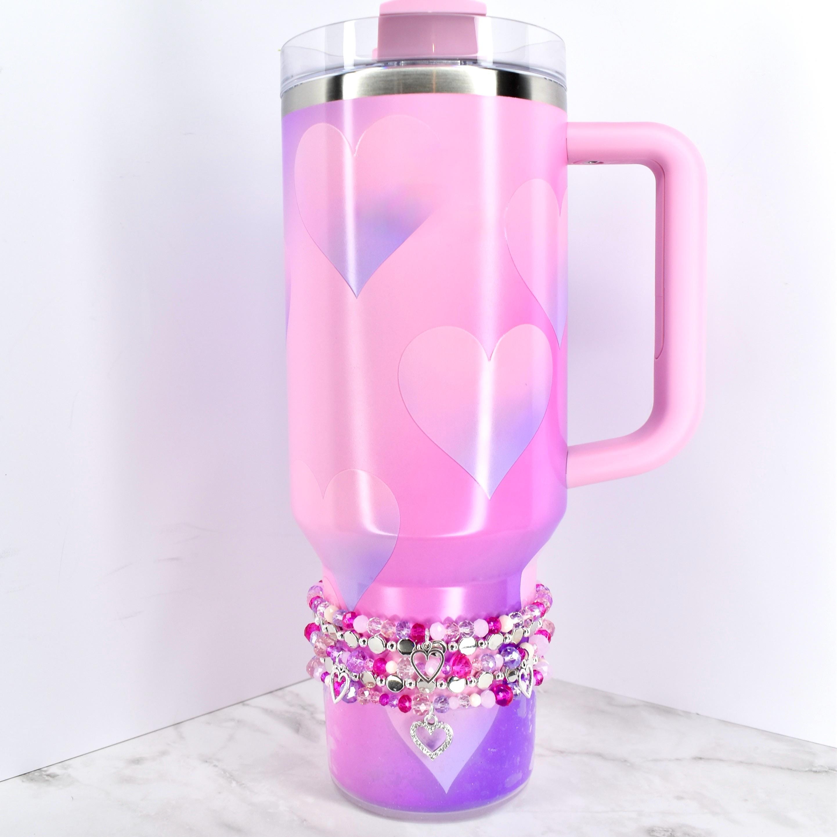 A pink insulated tumbler with a handle and a clear lid, decorated with large heart shapes in a lighter pink and purple gradient. A stack of beaded bracelets with heart charms adorns the base of the tumbler.