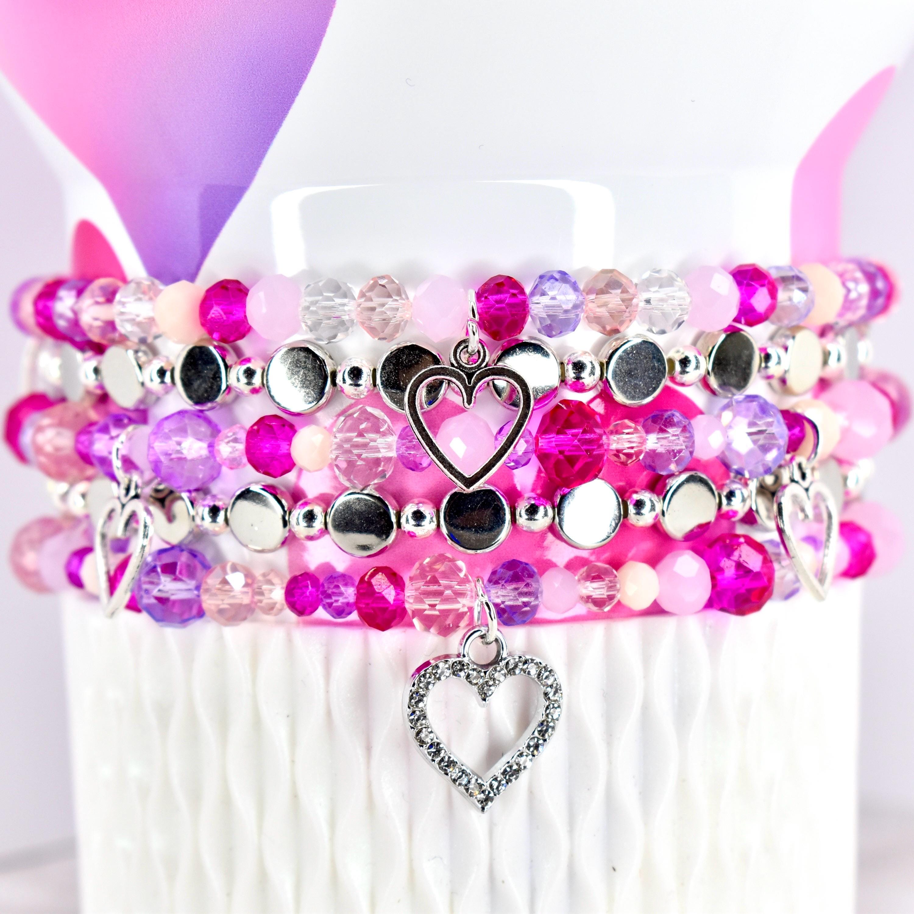 A stack of beaded bracelets in shades of pink, purple, and clear. Each bracelet features a mix of round and faceted beads, with silver-toned accents and heart-shaped charms. The bracelets are displayed on a white surface.