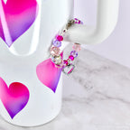 A beaded bracelet with pink, purple, and clear beads, and a silver heart charm. The bracelet is draped over the handle of a white mug decorated with pink and purple heart shapes.