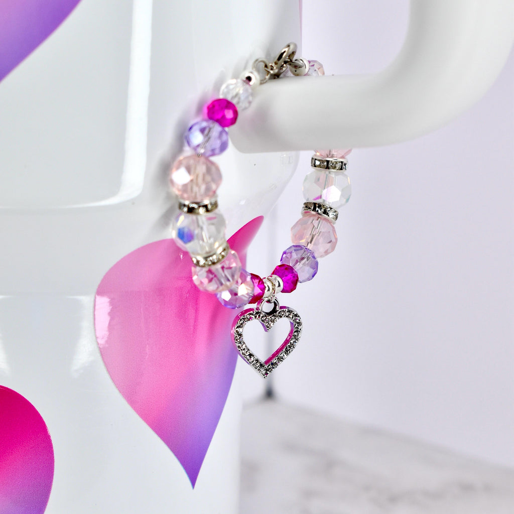 A beaded bracelet with a heart charm. The bracelet features a mix of pink, purple, and clear beads, with silver-toned accents. A silver heart charm, encrusted with small clear stones, dangles from the bracelet. The bracelet is displayed on a white background.