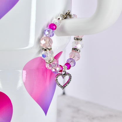 A beaded bracelet with a heart charm. The bracelet features a mix of pink, purple, and clear beads, with silver-toned accents. A silver heart charm, encrusted with small clear stones, dangles from the bracelet. The bracelet is displayed on a white background.