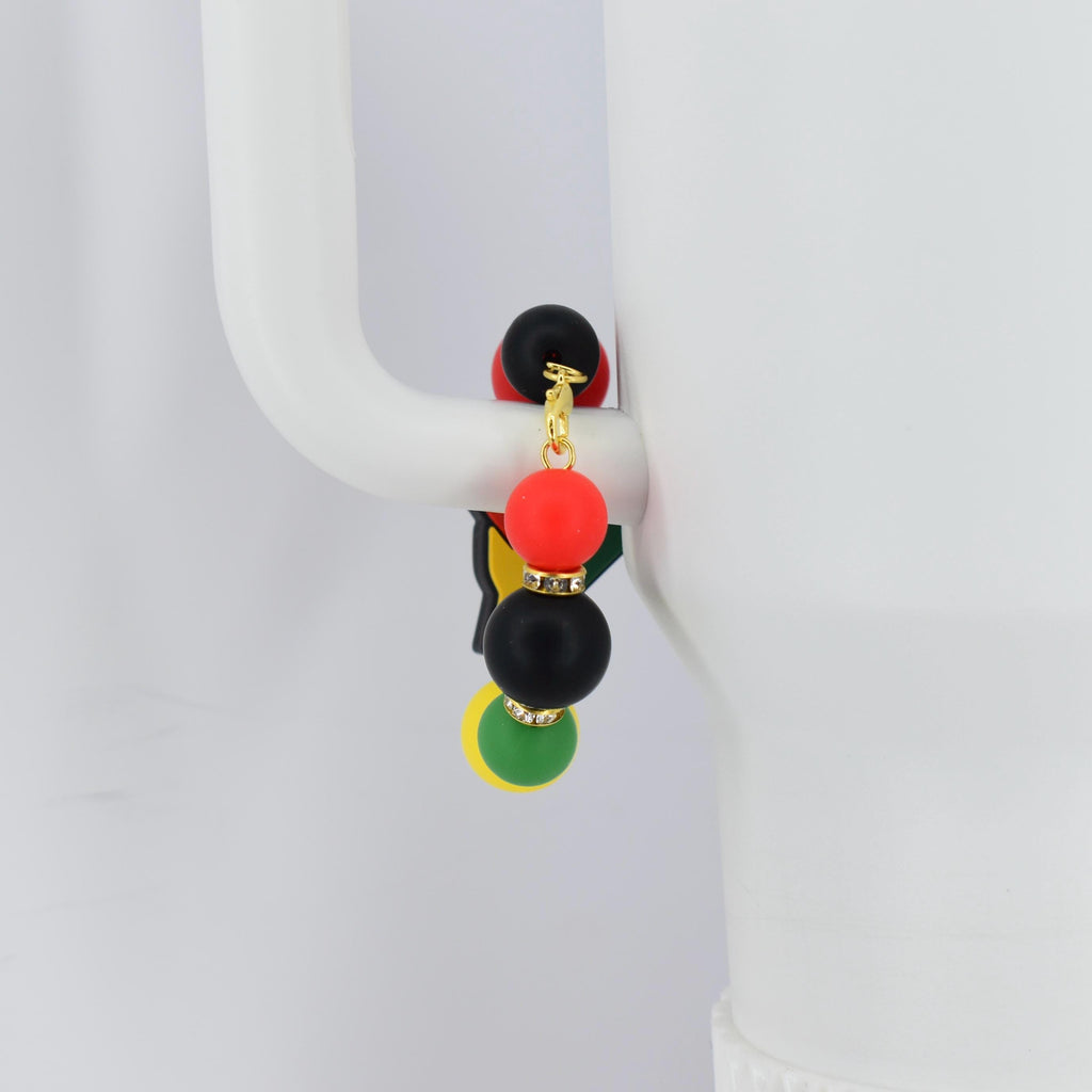 A tumbler bracelet with a gold-colored chain and colorful beads. The bracelet features a black, red, yellow, and green color scheme, with small rhinestones. The bracelet is hanging on a white mug handle.