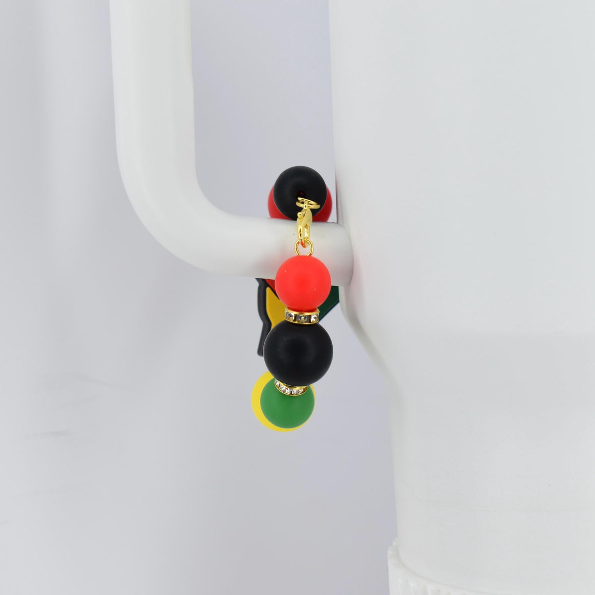 A tumbler bracelet with a gold-colored chain and colorful beads. The bracelet features a black, red, yellow, and green color scheme, with small rhinestones. The bracelet is hanging on a white mug handle.