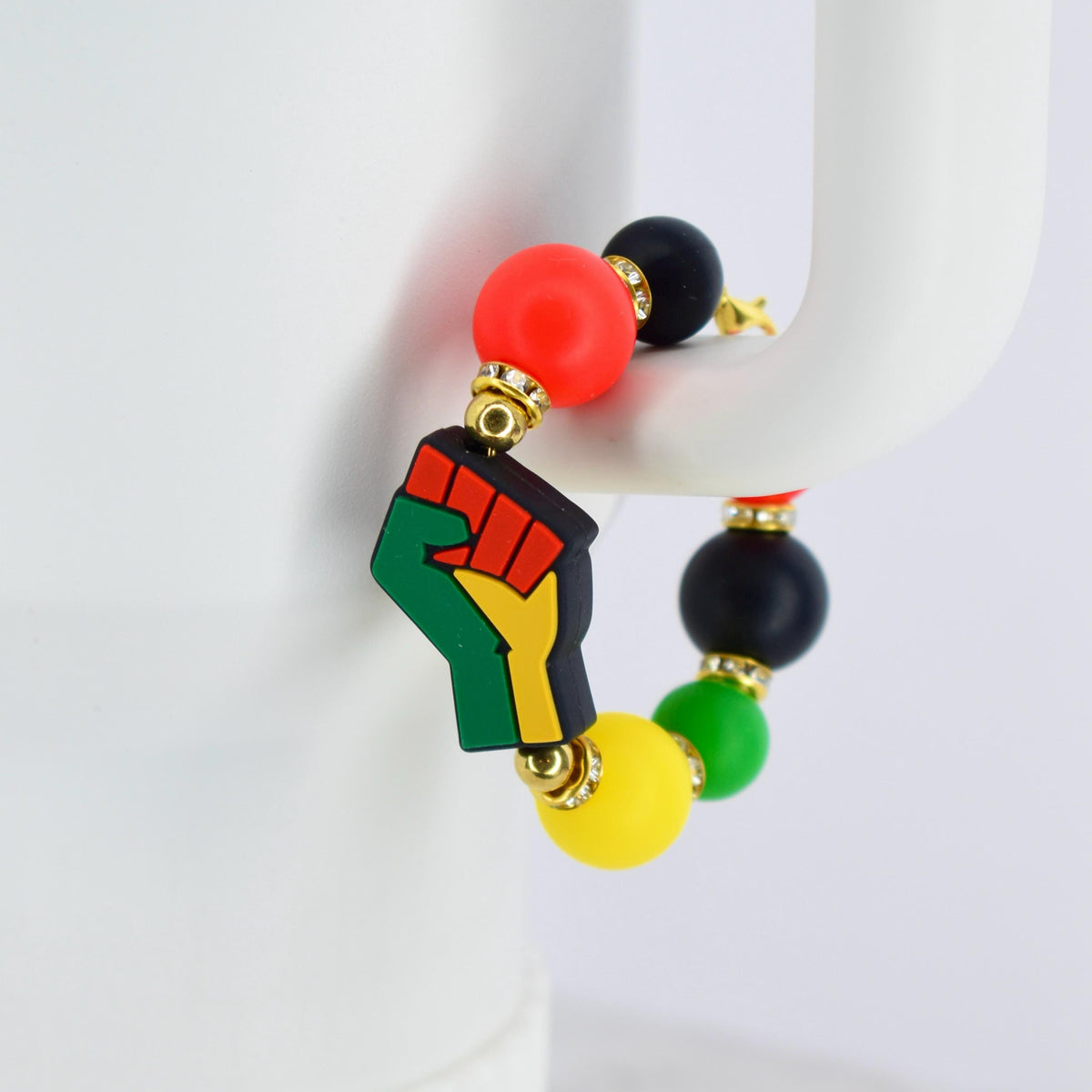 A beaded bracelet featuring a raised fist charm in black, green, red, and yellow. The bracelet includes red, black, green, and yellow beads, with gold-colored accents. The bracelet is displayed against a white background.