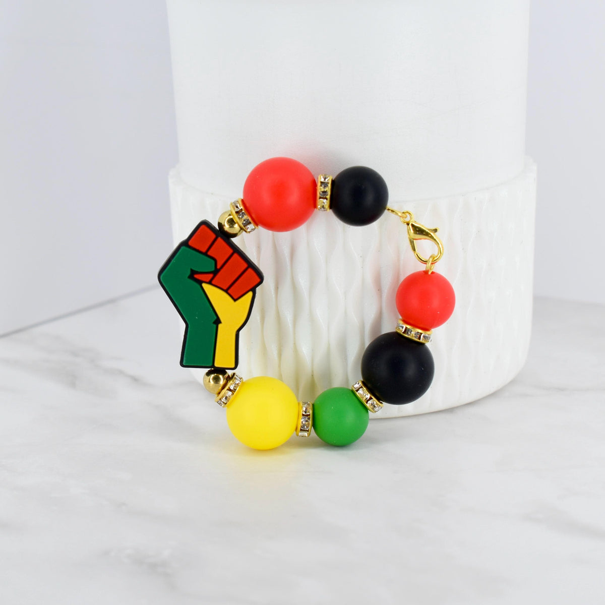 A beaded bracelet featuring a raised fist emblem in green, red, yellow, and black. The bracelet includes red, black, yellow, and green beads, with gold-colored accents. The bracelet is a symbol of solidarity.