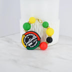 A beaded bracelet with Juneteenth-themed colors and a central charm. The bracelet features green, yellow, red, and black beads, with gold accents. The charm reads "Juneteenth Since 1865 Black History" in a circular design.