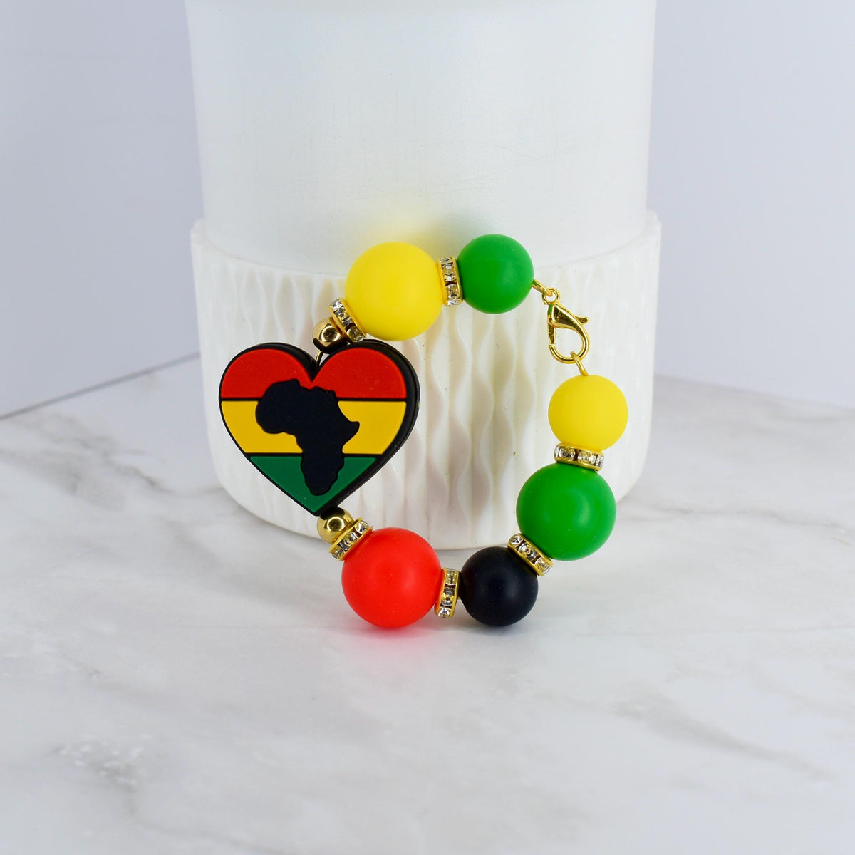 A beaded bracelet featuring a heart-shaped charm with the African continent silhouette in black, yellow, red, and green. The bracelet includes yellow, green, red, and black beads, connected by gold-colored accents and a clasp.