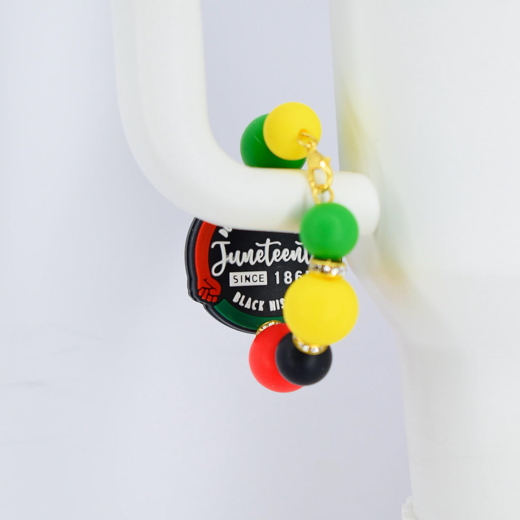 A pair of Juneteenth earrings with a gold chain and colorful beads. The earrings feature green, yellow, red, and black beads. The Juneteenth logo is visible on a black and green tag.