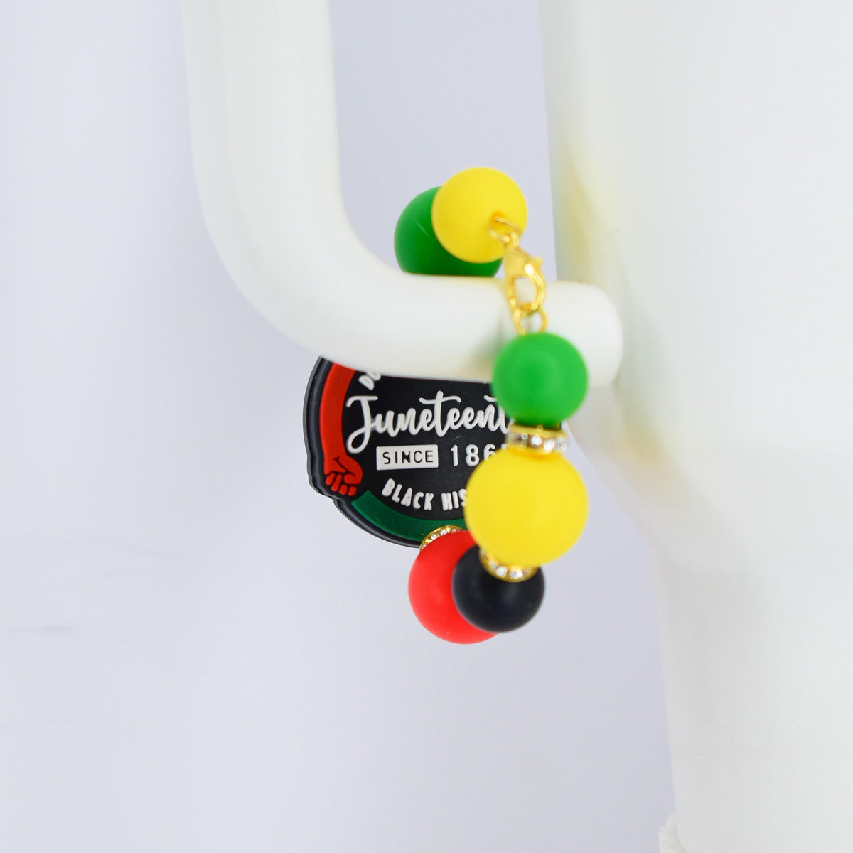A pair of Juneteenth earrings with a gold chain and colorful beads. The earrings feature green, yellow, red, and black beads. The Juneteenth logo is visible on a black and green tag.