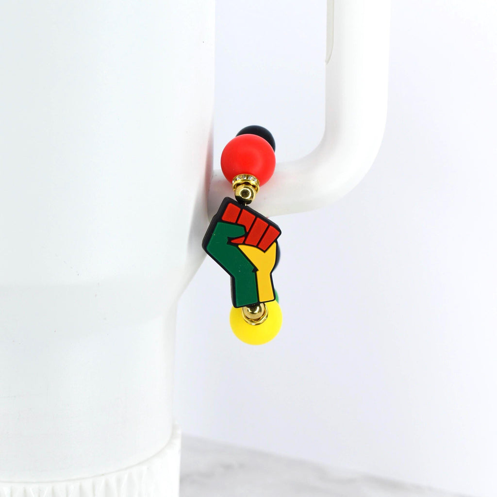 A tumbler handle charm featuring a fist design in green, yellow, and red, with a red and black bead at the top and a yellow bead at the bottom. The charm is hanging on a white surface.