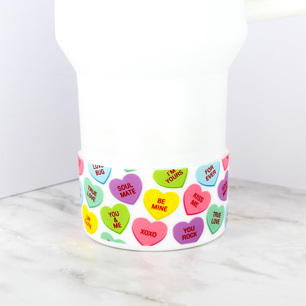 A white tumbler with a decorative band featuring colorful conversation heart designs. The hearts display phrases like "Be Mine," "Kiss Me," and "Soul Mate" in various colors, creating a festive and romantic theme.