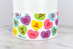 A white cylindrical container with a band of colorful conversation heart candies. The hearts have phrases like "Be Mine," "Kiss Me," and "Soul Mate." The background is a light gray surface.