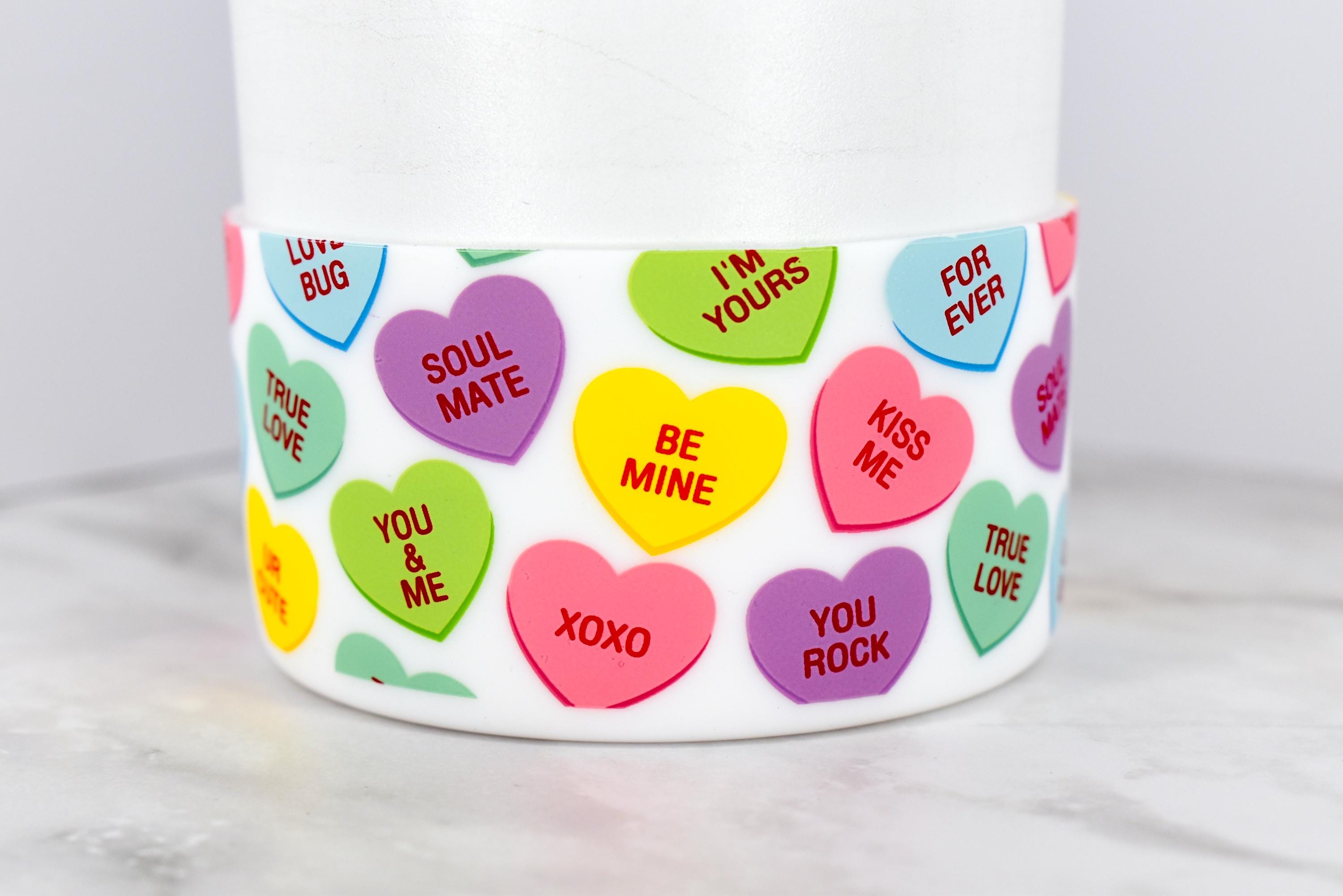 A white cylindrical container with a band of colorful conversation heart candies. The hearts have phrases like "Be Mine," "Kiss Me," and "Soul Mate." The background is a light gray surface.