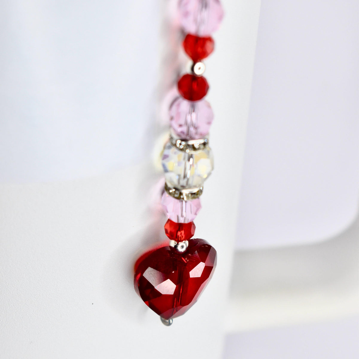 A dangling beaded ornament featuring a faceted red heart pendant. The ornament has a vertical design with alternating pink and red beads, along with clear and silver accents. The heart is the focal point, with a shiny, reflective surface.
