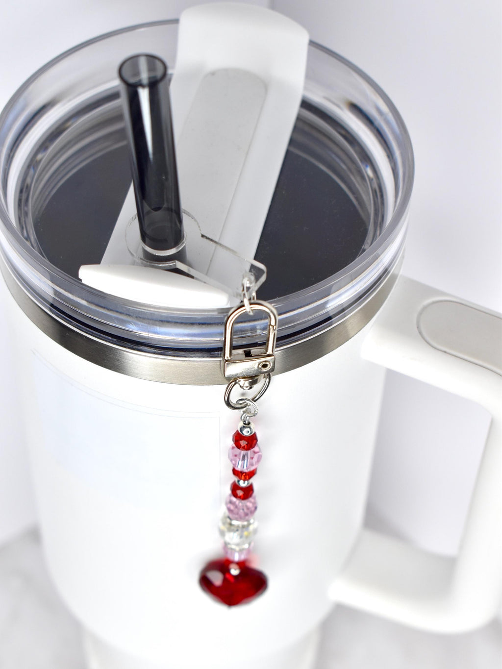 A white insulated tumbler with a clear lid and black straw. A decorative charm with red and pink beads and a red heart pendant hangs from the lid. The tumbler has a silver rim and a white handle.