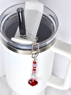 A white insulated tumbler with a clear lid and black straw. A decorative charm with red and pink beads and a red heart pendant hangs from the lid. The tumbler has a silver rim and a white handle.