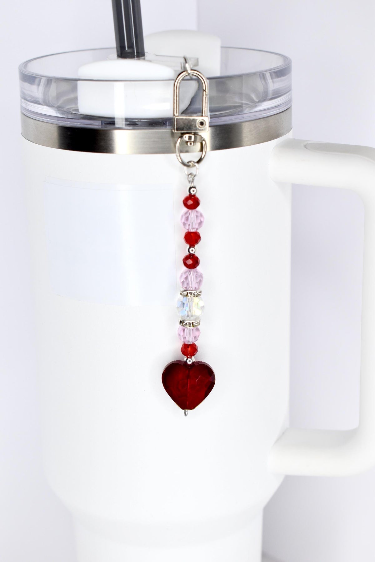 A beaded straw charm with a silver clasp attached to a white tumbler. The charm features a series of red, pink, and clear beads, culminating in a heart-shaped pendant. The tumbler has a clear lid and a black straw.