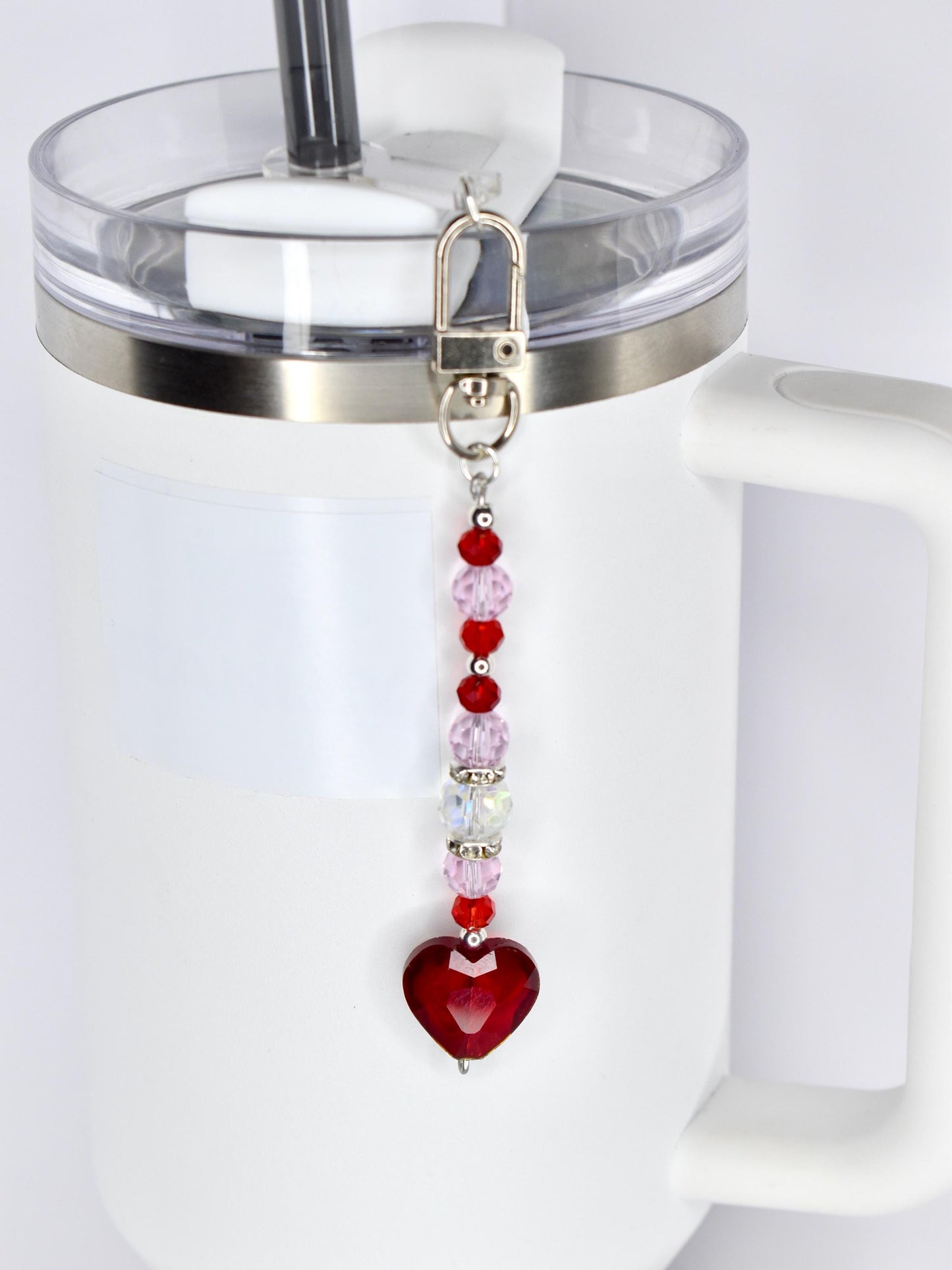 A red heart-shaped charm with a silver clasp and a beaded chain of red, pink, and clear beads. The charm is attached to a white tumbler with a clear lid and a silver rim.
