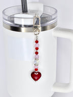 A red heart-shaped charm with a silver clasp and a beaded chain of red, pink, and clear beads. The charm is attached to a white tumbler with a clear lid and a silver rim.