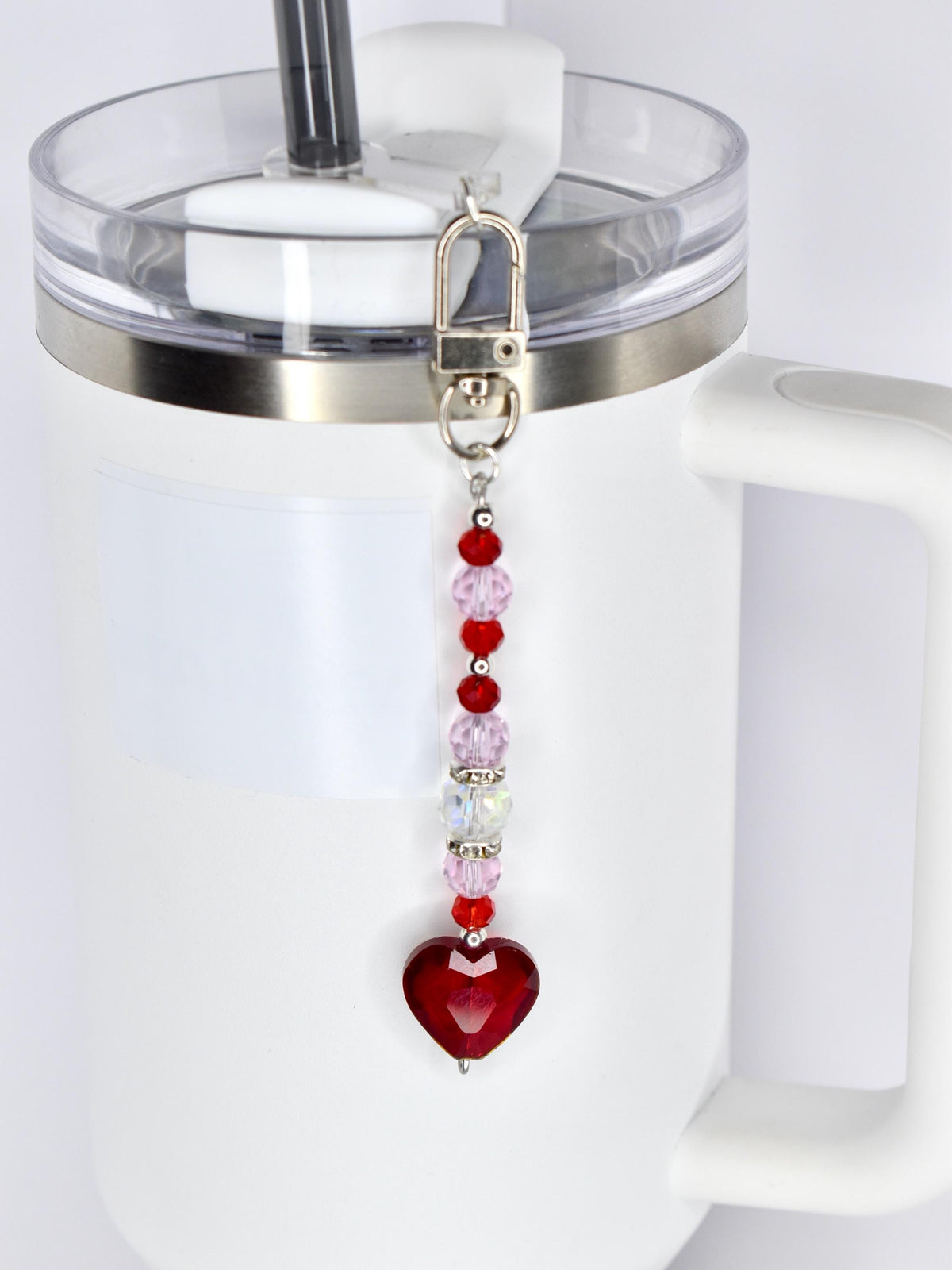 A red heart-shaped charm with a silver clasp and a beaded chain of red, pink, and clear beads. The charm is attached to a white tumbler with a clear lid and a silver rim.