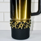 A black and gold insulated tumbler with a black handle. The tumbler is decorated with multiple strands of black and gold beads. The bottom of the tumbler has a black, textured, non-slip base.