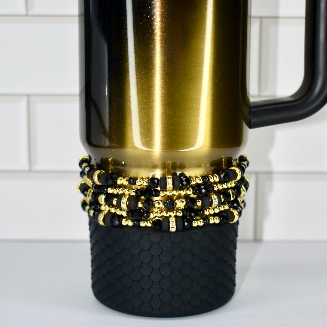 A black and gold insulated tumbler with a black handle. The tumbler is decorated with multiple strands of black and gold beads. The bottom of the tumbler has a black, textured, non-slip base.