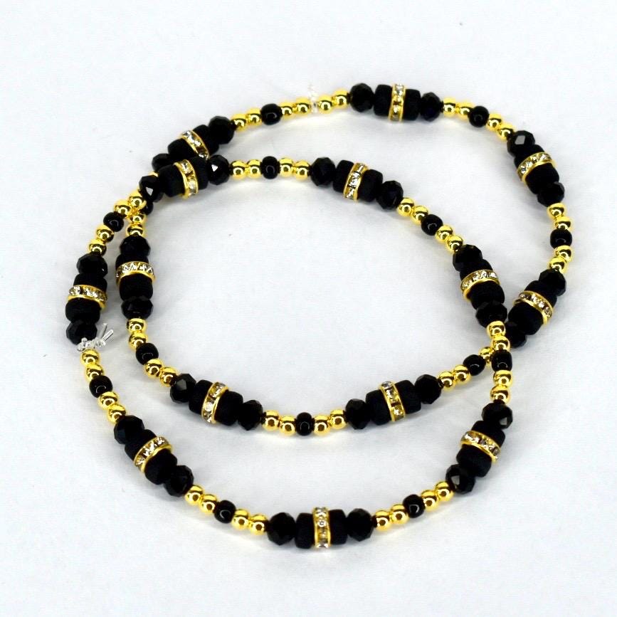 A set of two black beaded bracelets with gold-colored accents and rhinestone-studded spacers. The bracelets feature small, round gold beads and larger, faceted black beads. The design is symmetrical, with the beads arranged in a repeating pattern.