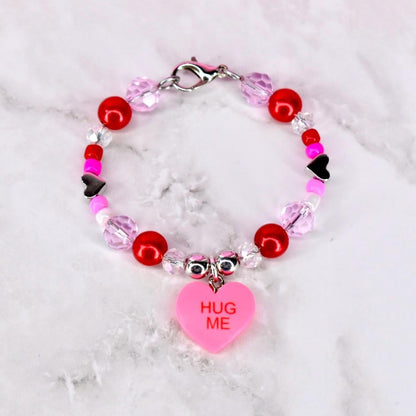 A beaded bracelet with red, pink, and clear beads. A pink heart charm with the words "HUG ME" hangs from the bracelet. The bracelet has a silver clasp.