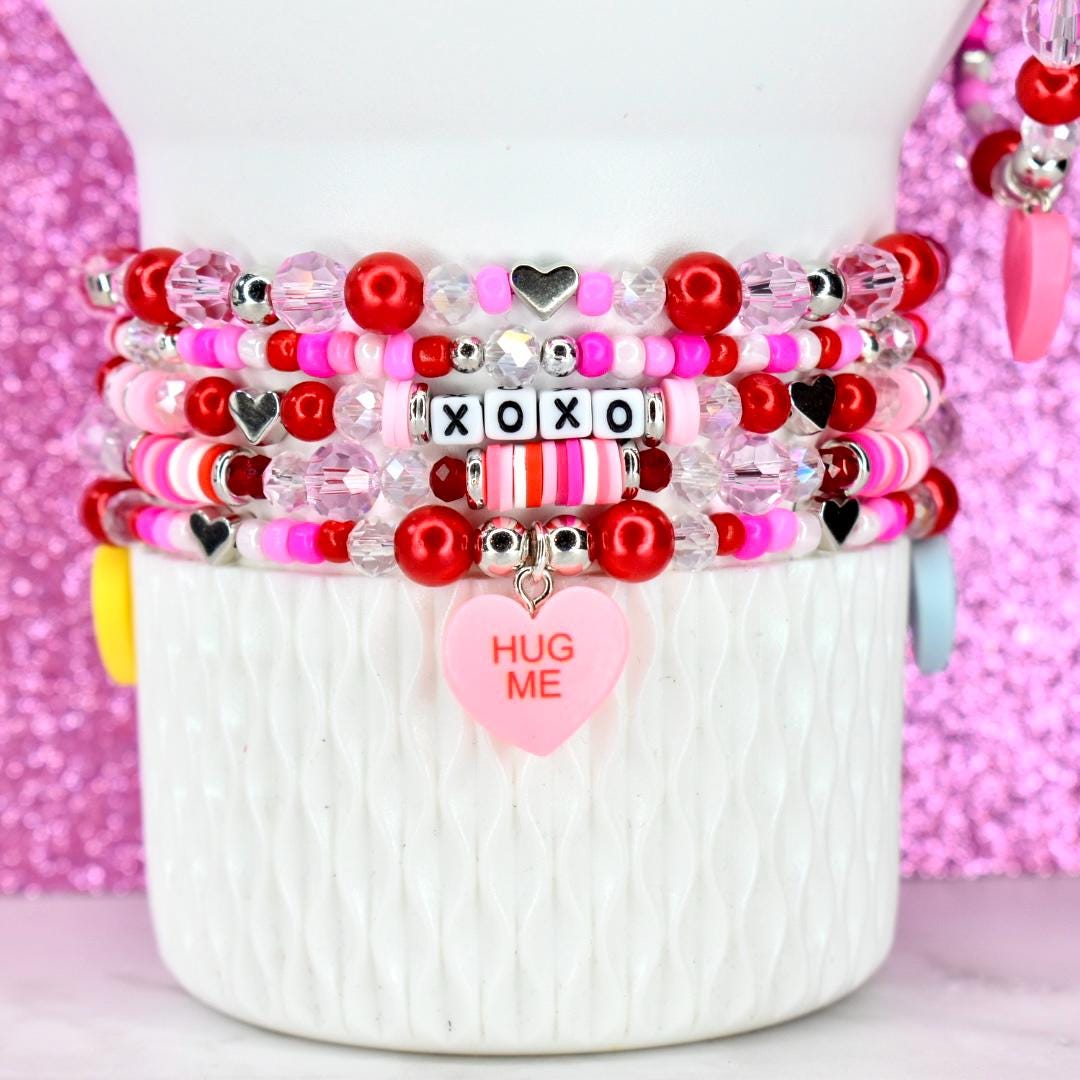 Stack of beaded bracelets in shades of red, pink, and white, with heart and XOXO accents. One bracelet features a pink heart charm that says "HUG ME". The bracelets are displayed on a white, textured surface.