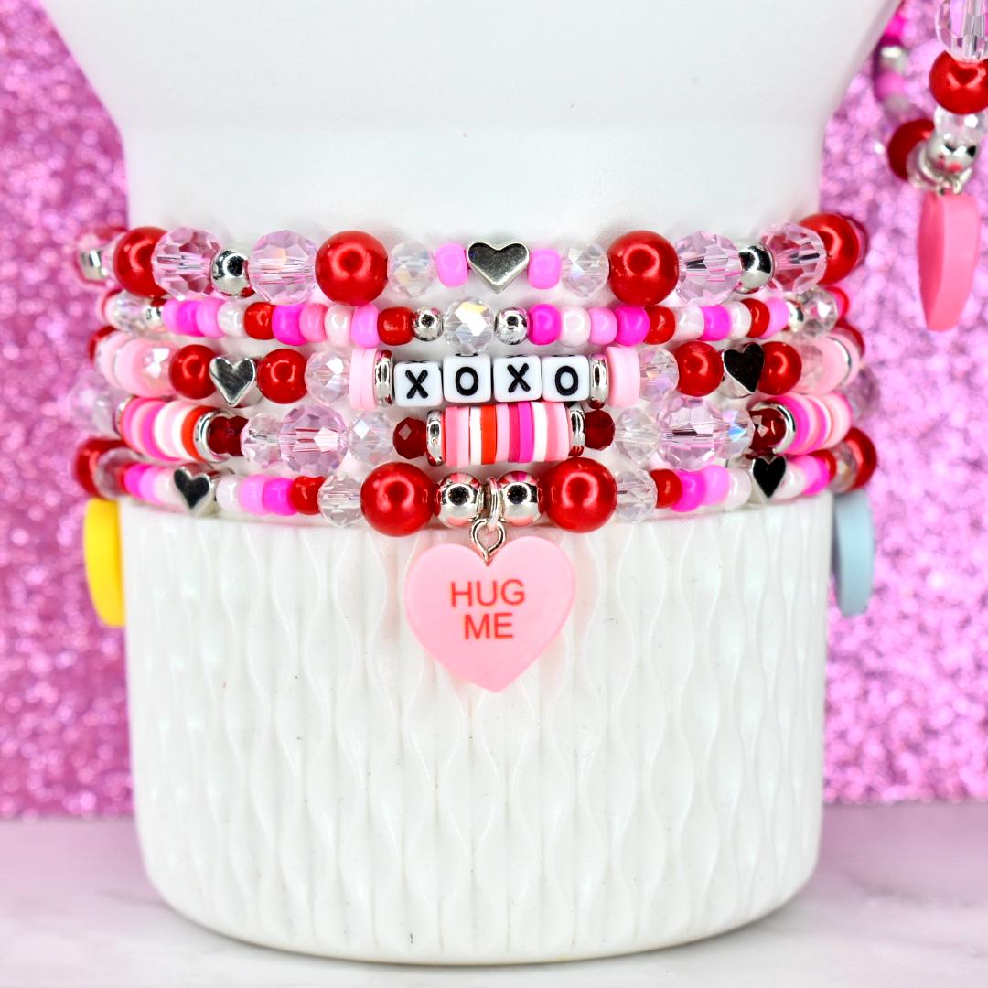 Stack of beaded bracelets in shades of red, pink, and white, with heart and XOXO accents. One bracelet features a pink heart charm that says "HUG ME". The bracelets are displayed on a white, textured surface.